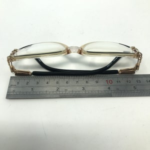 Puede incluir: Gafas con montura marr&oacute;n claro y lentes transparentes. Las patillas tienen detalles dorados y el puente es negro. Una regla debajo de las gafas mide aproximadamente 13 cm de largo.