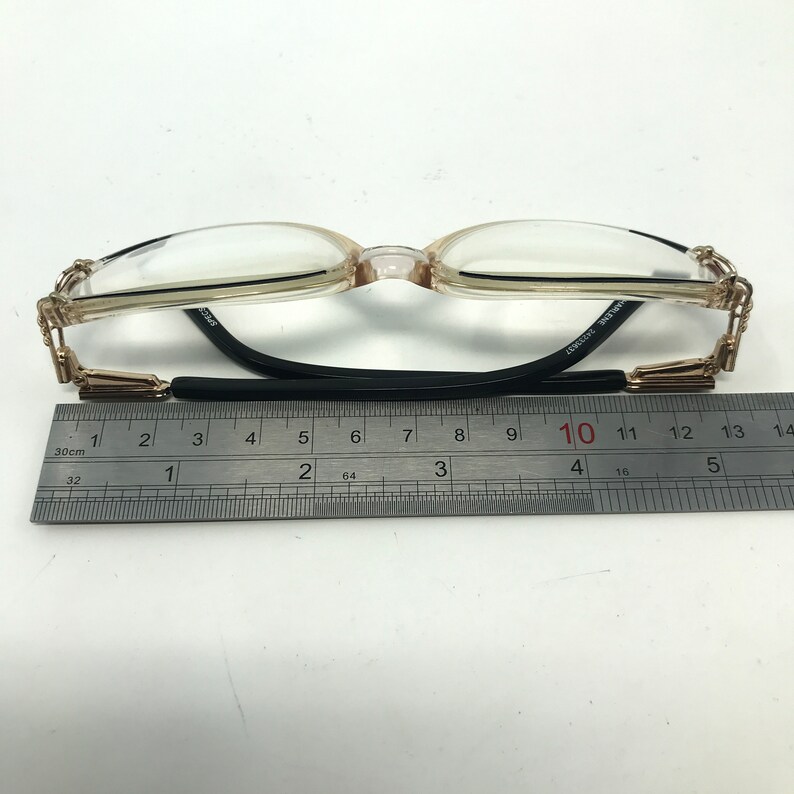 Puede incluir: Gafas con montura marr&oacute;n claro y lentes transparentes. Las patillas tienen detalles dorados y el puente es negro. Una regla debajo de las gafas mide aproximadamente 13 cm de largo.