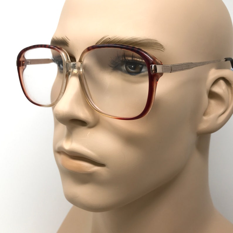 K&ouml;nnte beinhalten: Eine Brille in Braun und Gold mit klaren Gl&auml;sern. Die Brille hat eine quadratische Form und einen dicken Rahmen.