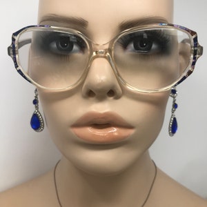 Puede incluir: Gafas con montura transparente de forma rectangular con detalles decorativos en las patillas. La imagen tambi&eacute;n presenta un par de pendientes colgantes con gemas azules en forma de l&aacute;grima y metal plateado.