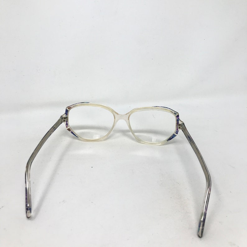 Puede incluir: Gafas transparentes con montura de color claro y lentes rectangulares. Las patillas son plateadas con un patr&oacute;n sutil. La montura tiene un patr&oacute;n decorativo en tonos azules, morados y blancos cerca de las bisagras.