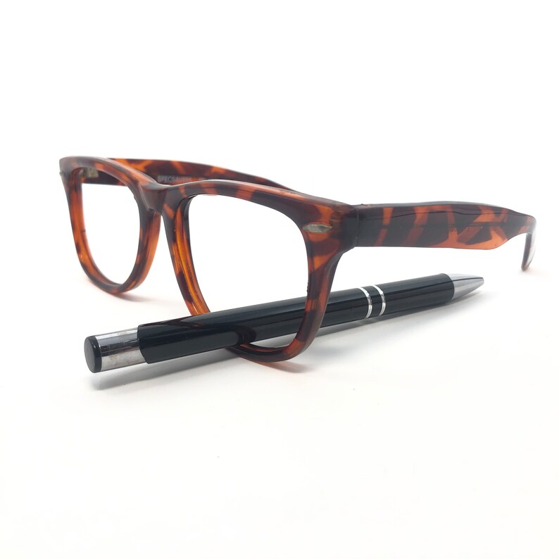K&ouml;nnte beinhalten: Eine Brille mit Schildpattmuster und ein schwarzer Stift. Die Brille hat einen klassischen rechteckigen Rahmen. Der Stift ist schwarz mit silbernen Details, vor wei&szlig;em Hintergrund.