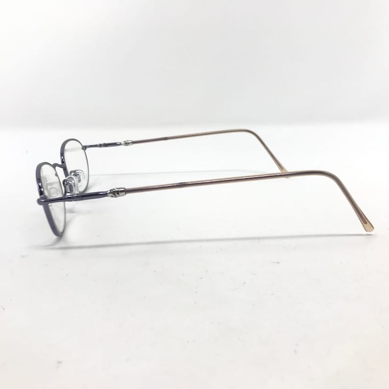 May include: A pair of eyeglasses with a thin, oval-shaped frame. The frame is a dark, muted color, with slender, light brown temples. The lenses are clear, and the glasses are set against a white background.