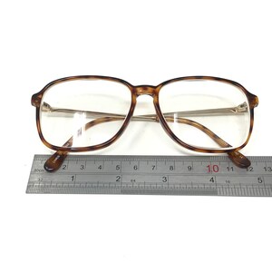 K&ouml;nnte beinhalten: Tortoiseshell-Brille mit goldenen Metallb&uuml;geln und klaren Gl&auml;sern. Die Brille wird mit einem Lineal gemessen.