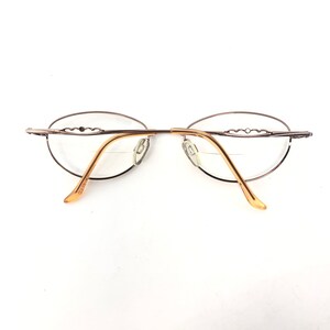 May include: Oval-shaped eyeglasses with a thin, silver-colored metal frame and gold-toned temples. The lenses are clear, and the design features a decorative element on the upper frame. The glasses are set against a white background.