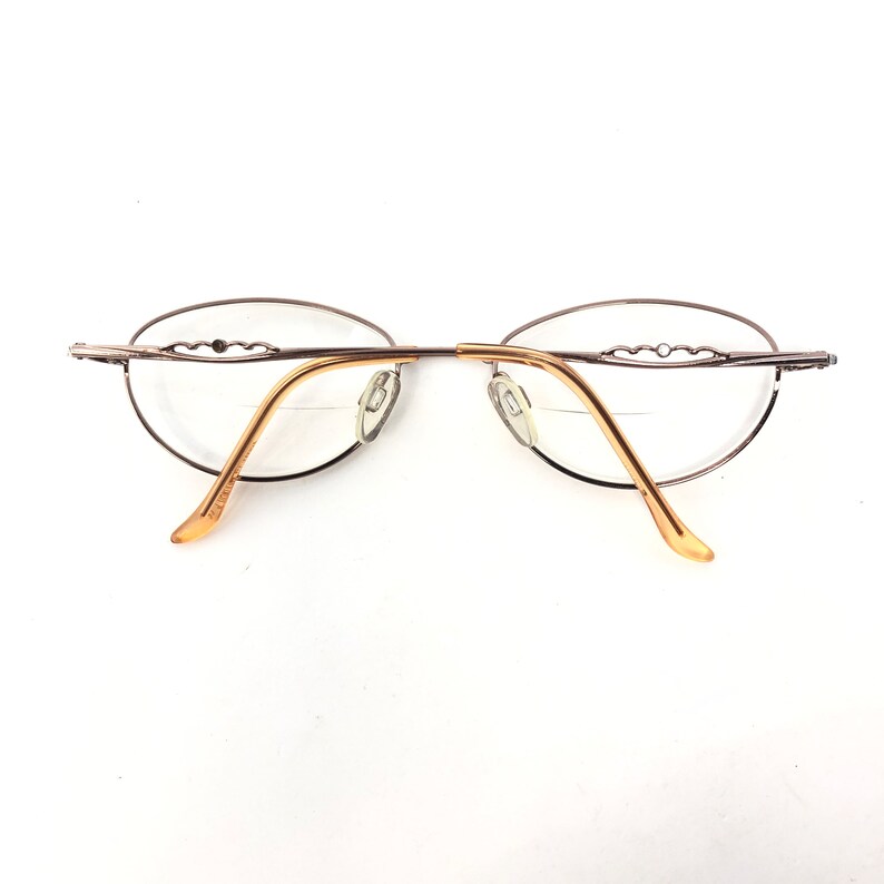 May include: Oval-shaped eyeglasses with a thin, silver-colored metal frame and gold-toned temples. The lenses are clear, and the design features a decorative element on the upper frame. The glasses are set against a white background.