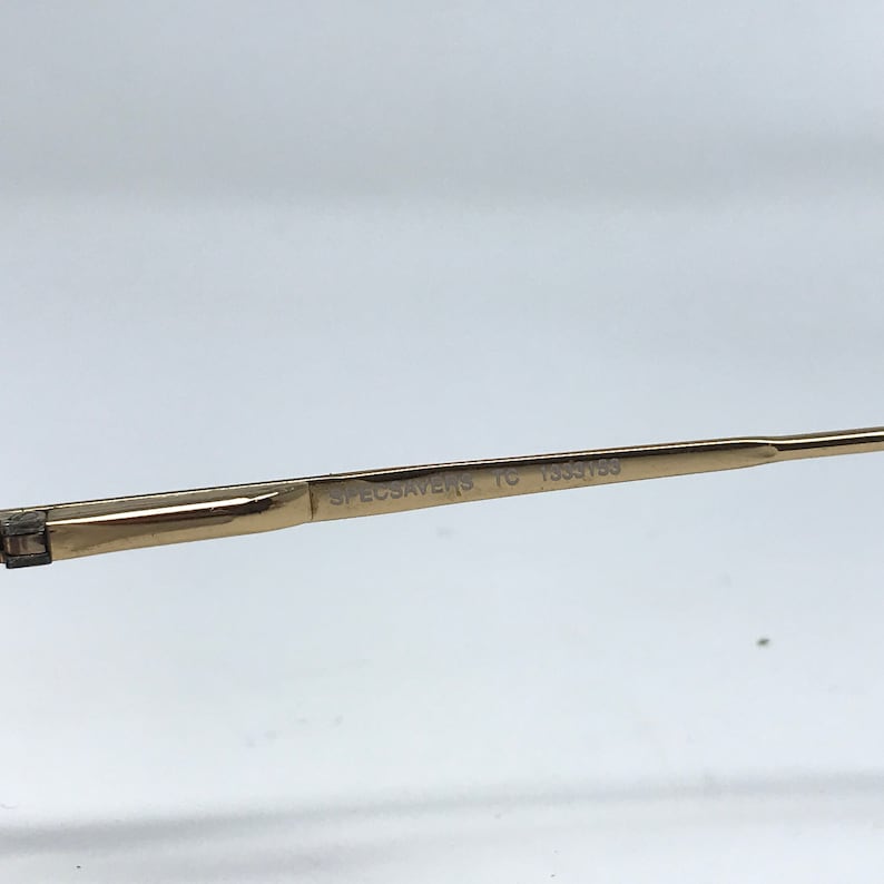 May include: Close-up of a gold-colored eyeglass frame arm. The text "SPECSAVERS TC 1333153" is printed on the arm. The frame is set against a white background.