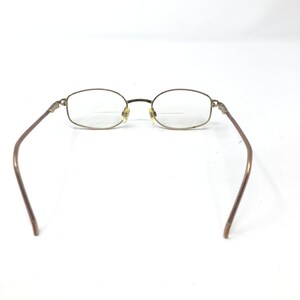 May include: Eyeglasses with a gold-toned metal frame and oval lenses. The temples are a darker brown colour. The glasses are set against a white background.