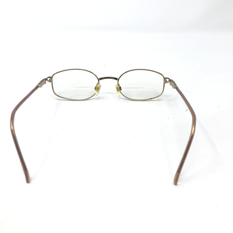May include: Eyeglasses with a gold-toned metal frame and oval lenses. The temples are a darker brown colour. The glasses are set against a white background.
