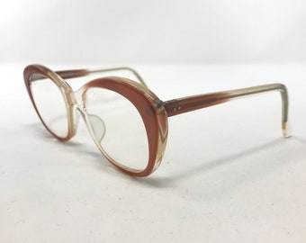 May include: Vintage cat-eye eyeglasses. The frames are a gradient of brown and clear plastic. The lenses are clear, and the temples are a light brown colour. The glasses are set against a white background.