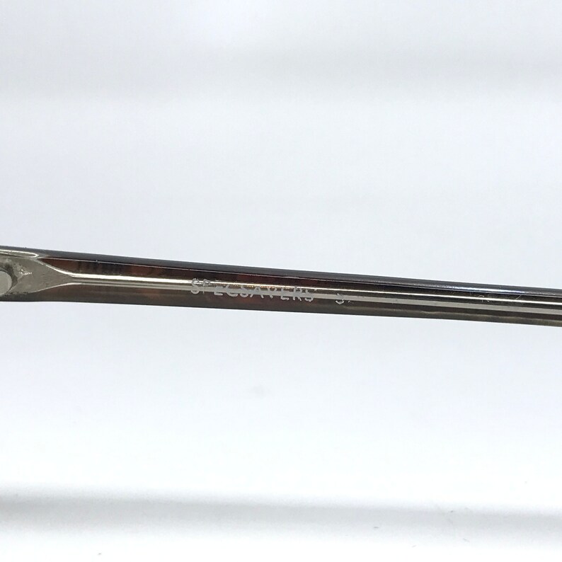 May include: Close-up of a portion of eyeglasses with the text "SPECSAVERS" printed on the temple. The frame is a dark metallic color, and the background is plain white.