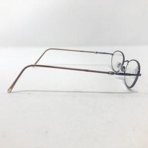 May include: A pair of eyeglasses with a thin, dark blue frame and oval lenses. The temples are a light brown color. The glasses are set against a white background.