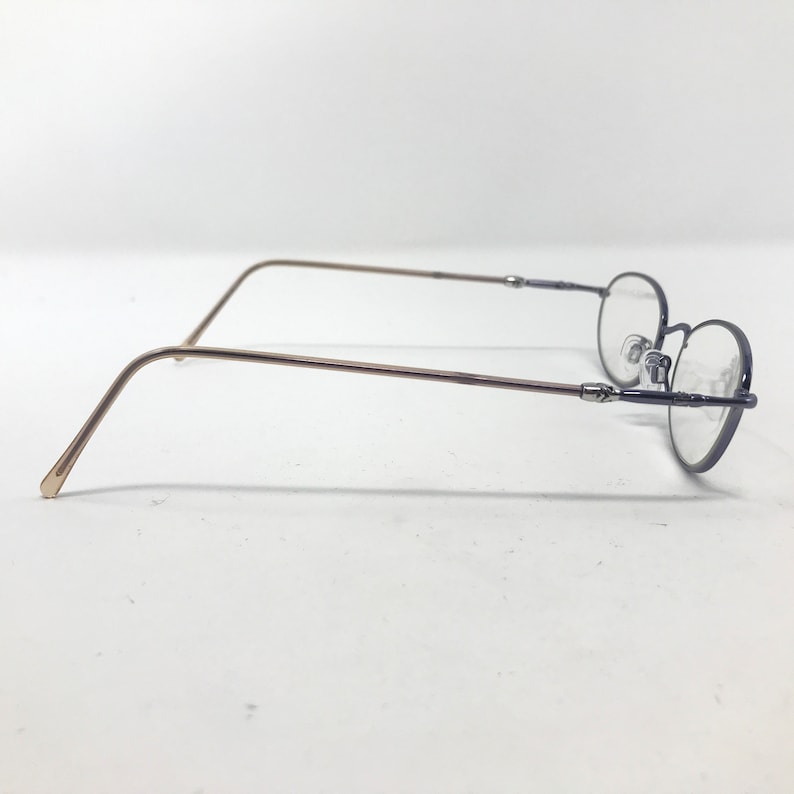 May include: A pair of eyeglasses with a thin, dark blue frame and oval lenses. The temples are a light brown color. The glasses are set against a white background.