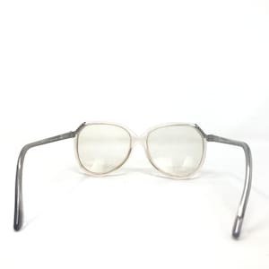 May include: Clear-framed eyeglasses with gray temples. The lenses are large and oval-shaped. The temples have text on them. The glasses are set against a white background.
