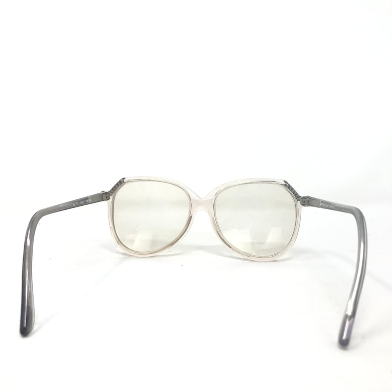 May include: Clear-framed eyeglasses with gray temples. The lenses are large and oval-shaped. The temples have text on them. The glasses are set against a white background.
