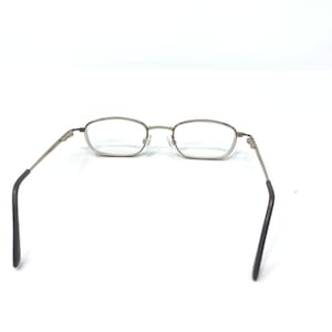 May include: Silver-framed eyeglasses with rectangular lenses. The glasses have a thin metal frame and dark gray temple arms. The lenses are clear, and the glasses are set against a white background.