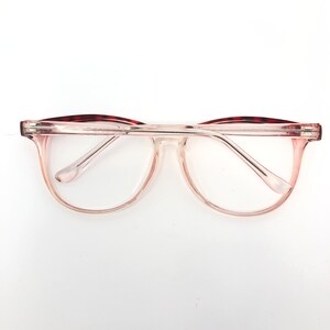 May include: Clear eyeglasses with a light pink gradient frame and clear lenses. The top of the frame has a dark red and brown tortoiseshell pattern. The glasses are set against a white background.