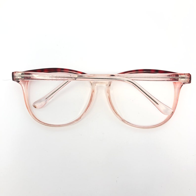 May include: Clear eyeglasses with a light pink gradient frame and clear lenses. The top of the frame has a dark red and brown tortoiseshell pattern. The glasses are set against a white background.
