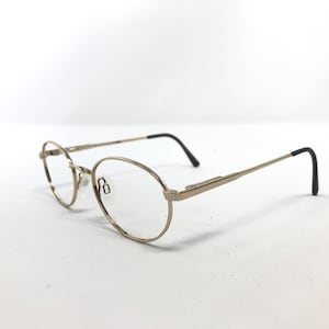 Pu&ograve; includere: Occhiali di forma ovale con montatura in metallo color oro e terminali neri delle aste. Le lenti sono trasparenti e i naselli sono trasparenti. Gli occhiali sono su uno sfondo bianco.