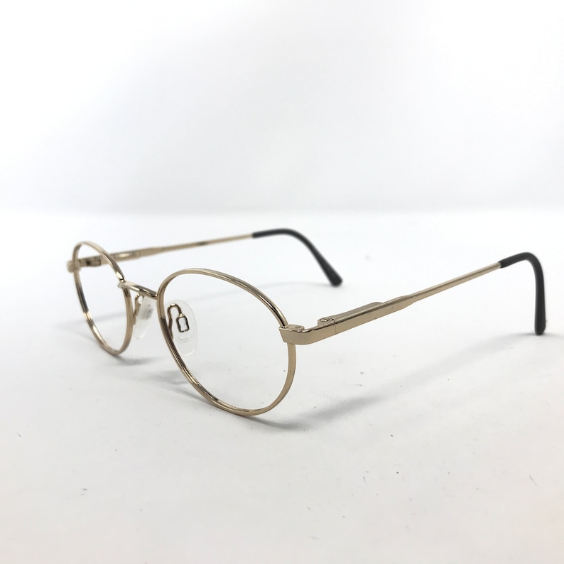 Pu&ograve; includere: Occhiali di forma ovale con montatura in metallo color oro e terminali neri delle aste. Le lenti sono trasparenti e i naselli sono trasparenti. Gli occhiali sono su uno sfondo bianco.