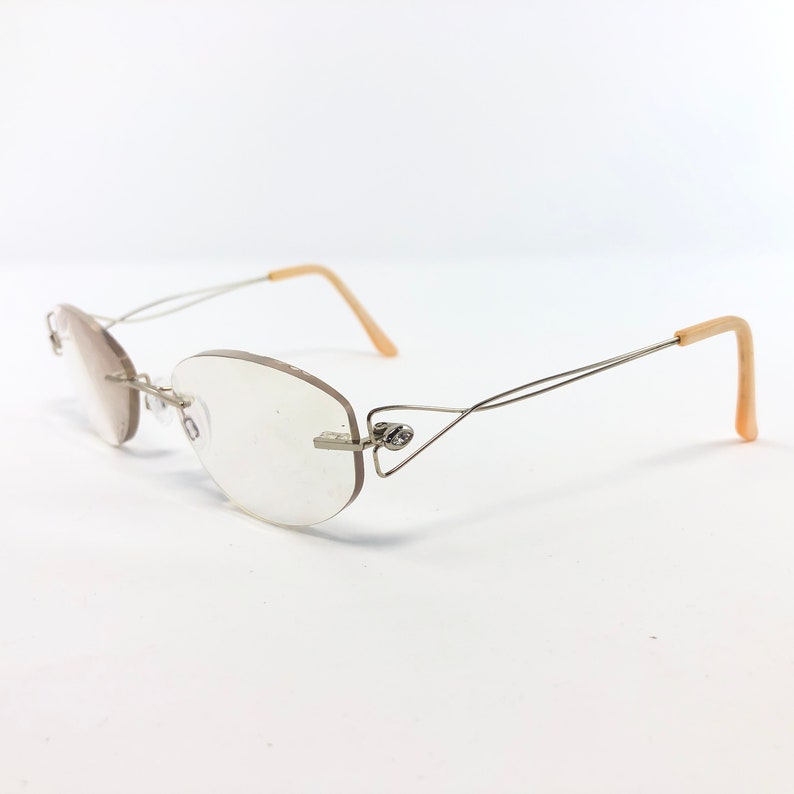 May include: Rimless eyeglasses with oval lenses and a silver metal frame. The temples are a light beige color. The glasses are set against a white background.