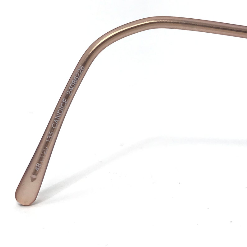 May include: Close-up of a pair of eyeglasses with a light brown frame. The text "48 12 135 CANDICE 24086226" is printed on the temple arm. The frame has a curved design.