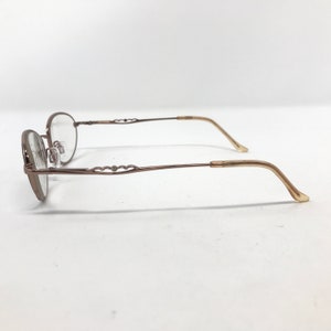 May include: Oval-shaped eyeglasses with a bronze-colored frame. The temples feature a decorative design, and the earpieces are a lighter shade of bronze. The lenses are clear, and the glasses are set against a white background.