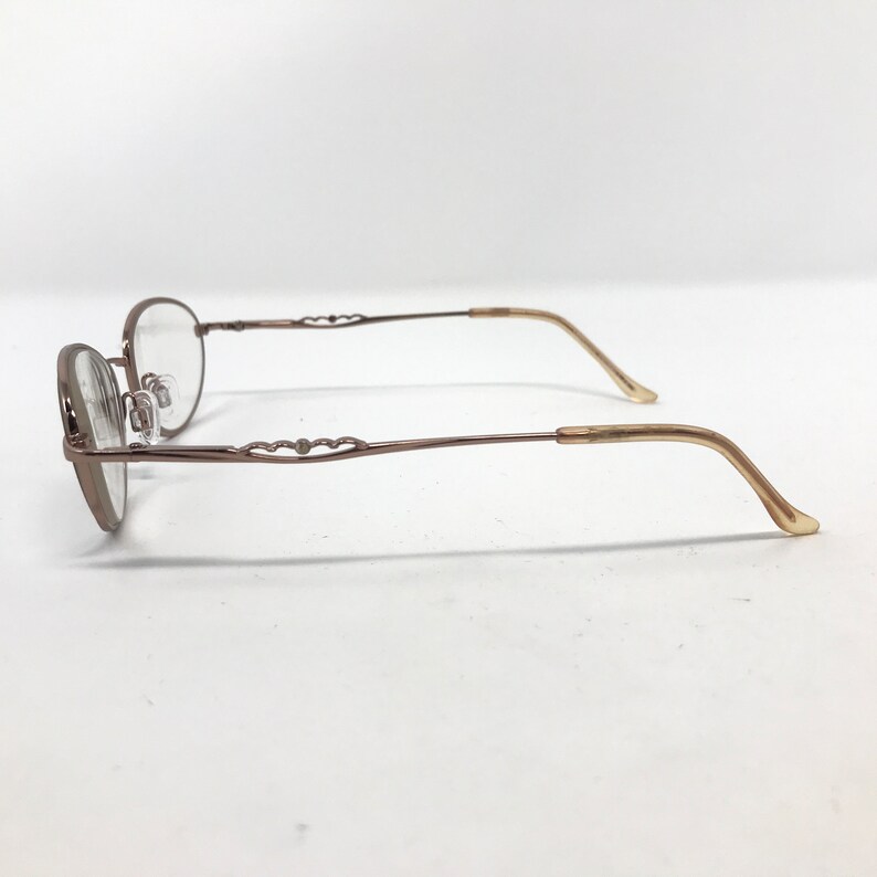 May include: Oval-shaped eyeglasses with a bronze-colored frame. The temples feature a decorative design, and the earpieces are a lighter shade of bronze. The lenses are clear, and the glasses are set against a white background.