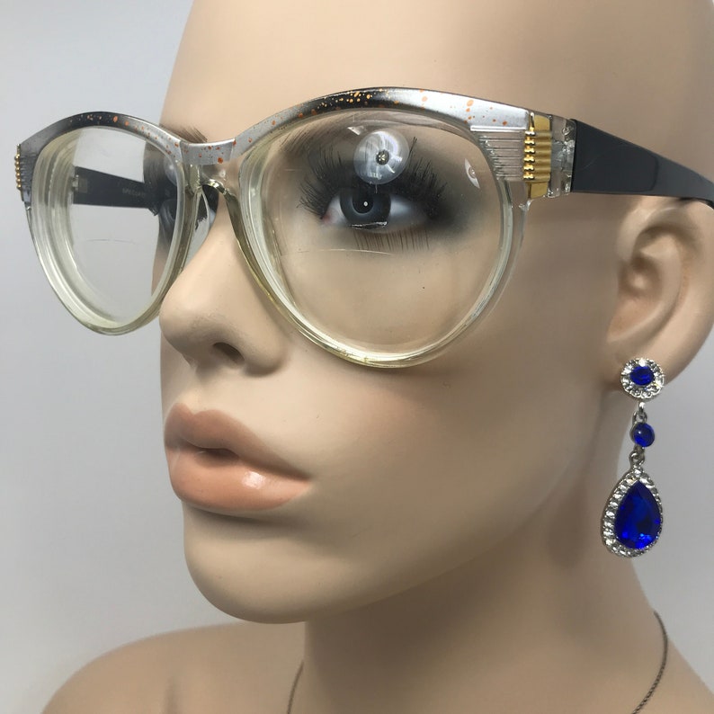 May include: Clear plastic framed glasses with gold accents and black arms. The glasses are on a mannequin head with blue teardrop earrings.
