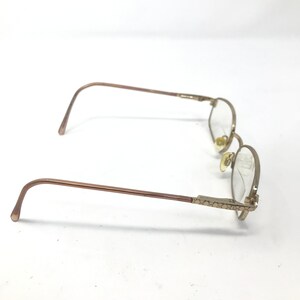 May include: Gold-toned eyeglasses with oval lenses. The frames have a thin, delicate design with decorative detailing on the temples. The glasses are set against a white background.
