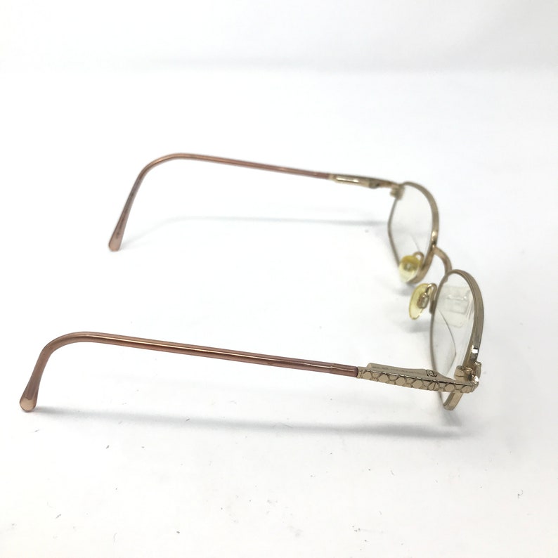 May include: Gold-toned eyeglasses with oval lenses. The frames have a thin, delicate design with decorative detailing on the temples. The glasses are set against a white background.