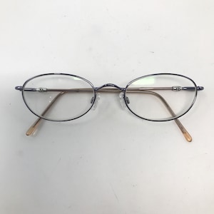 May include: Oval-shaped eyeglasses with a dark blue frame and clear lenses. The temples are a light brown color. The glasses have a classic, minimalist design.
