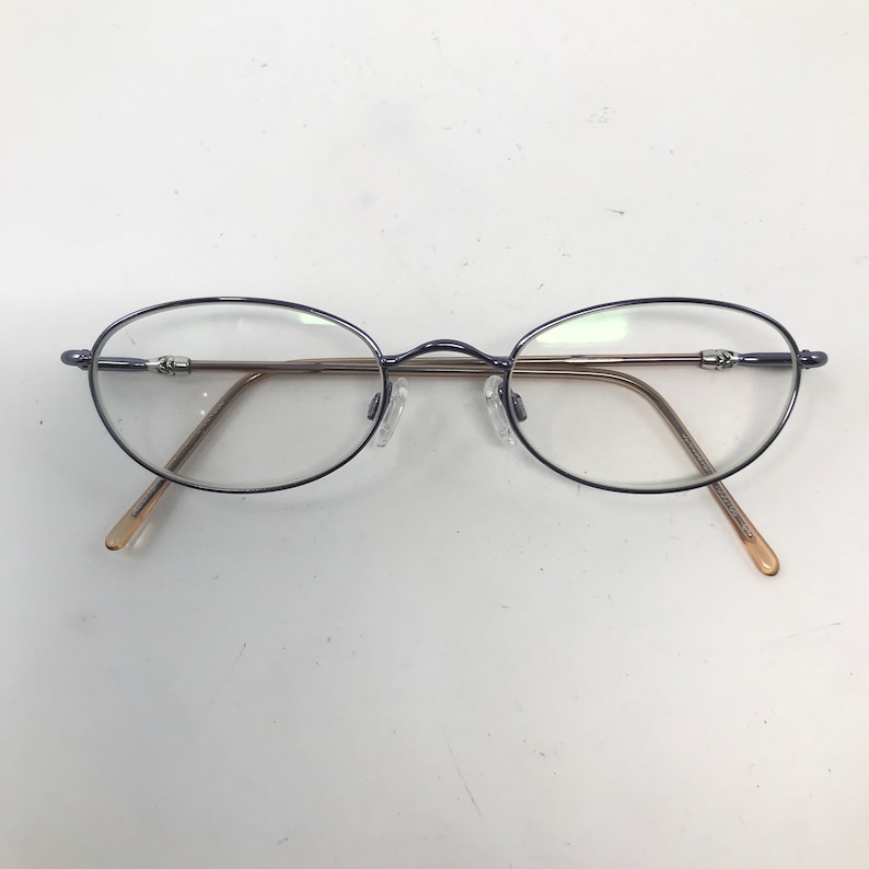 May include: Oval-shaped eyeglasses with a dark blue frame and clear lenses. The temples are a light brown color. The glasses have a classic, minimalist design.