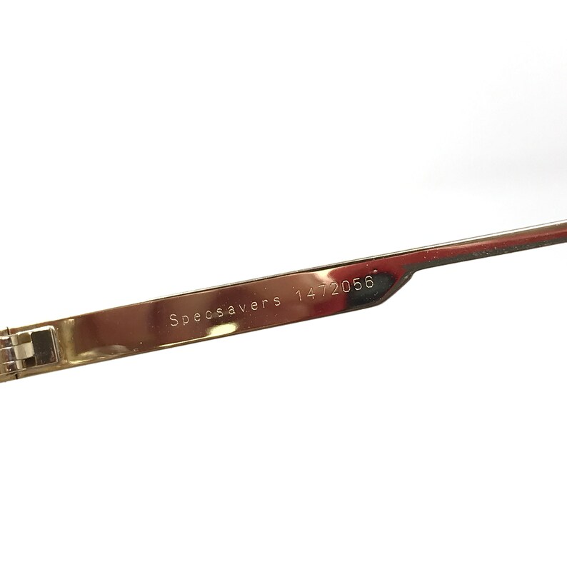 May include: Close-up of the arm of a pair of gold-tone glasses with the text "Specsavers 1472056" engraved on the metal.