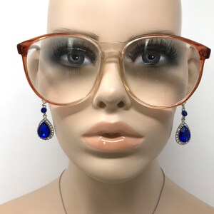 May include: Clear-lensed eyeglasses with a light brown frame are worn by a mannequin. The glasses are paired with dangling earrings featuring a large, blue teardrop-shaped gem surrounded by smaller clear stones. The mannequin has full eyelashes and glossy lips.