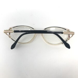 Puede incluir: Un par de gafas con montura transparente y patillas negras. Las gafas tienen un adorno dorado en las sienes. Las lentes son transparentes y las gafas est&aacute;n sobre un fondo blanco.