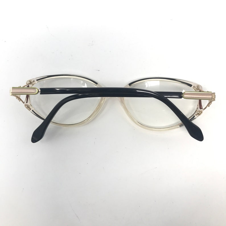 Puede incluir: Un par de gafas con montura transparente y patillas negras. Las gafas tienen un adorno dorado en las sienes. Las lentes son transparentes y las gafas est&aacute;n sobre un fondo blanco.