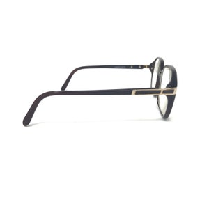 May include: Eyeglasses with a dark brown frame and temples. The lenses are clear, and the frame has a rectangular shape with gold-coloured accents. The glasses are viewed from the side against a white background.