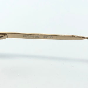 May include: Close-up of a gold-colored eyeglass arm with the text "SPECSAVERS 1853173 J" printed in black. The arm is thin and sleek, with a slight curve. The background is plain white.