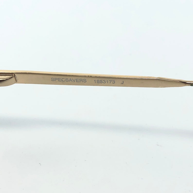 May include: Close-up of a gold-colored eyeglass arm with the text "SPECSAVERS 1853173 J" printed in black. The arm is thin and sleek, with a slight curve. The background is plain white.