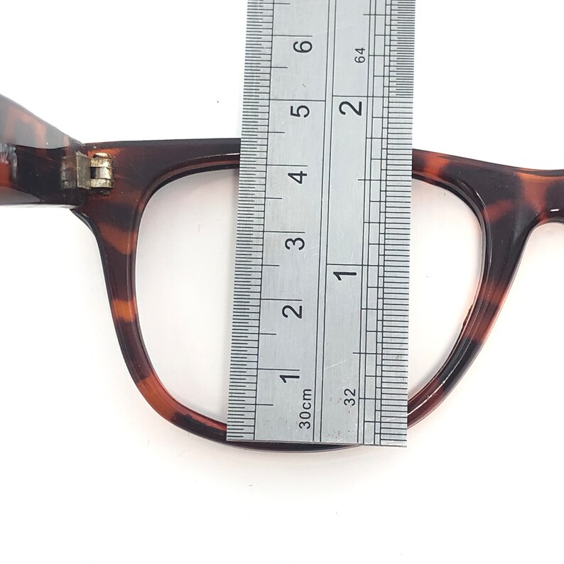 K&ouml;nnte beinhalten: Nahaufnahme einer Brille mit Schildpattmuster, bei der ein Lineal den Rahmen misst. Die Brille hat eine klassische rechteckige Form und ein braun-schwarzes Muster. Das Lineal zeigt, dass der Rahmen etwa 5 cm breit ist.