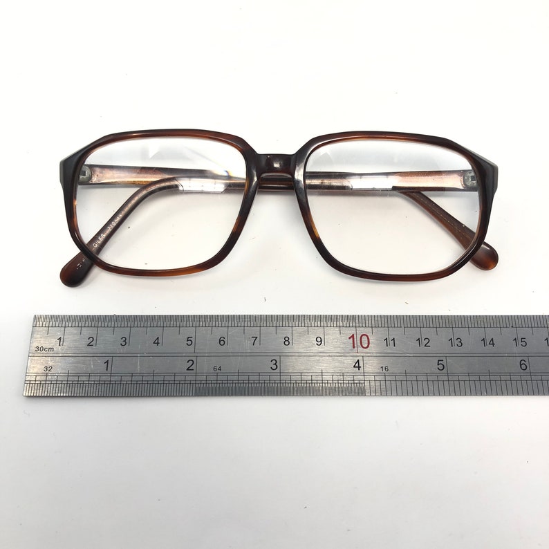 May include: Brown tortoiseshell eyeglasses with a rectangular frame. The glasses are placed next to a metal ruler, with measurements in inches. The frame has a classic, vintage design.