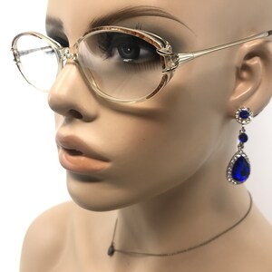 K&ouml;nnte beinhalten: Brille mit transparentem Rahmen und hellbraunen Akzenten an den oberen Ecken. Die Brille hat einen goldfarbenen Metallrahmen. H&auml;ngeohrringe mit blauen tropfenf&ouml;rmigen Edelsteinen und kleinen klaren Steinen sind ebenfalls zu sehen. Eine kleine Anh&auml;ngerkette ist ebenfalls vorhanden.