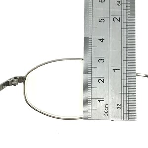 Op de afbeelding: Close-up van een zilverkleurige brilglazen lens met een liniaal. De lens meet ongeveer 3,8 cm in de breedte. Het montuur is dun en metaalachtig. De achtergrond is effen wit.