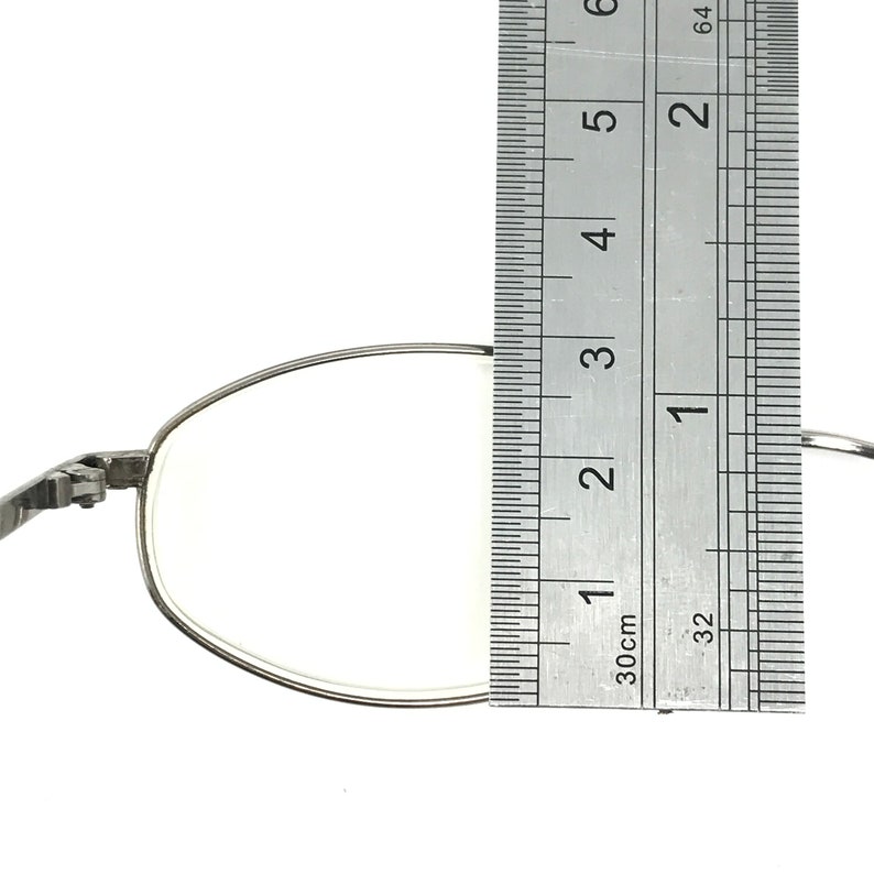 Op de afbeelding: Close-up van een zilverkleurige brilglazen lens met een liniaal. De lens meet ongeveer 3,8 cm in de breedte. Het montuur is dun en metaalachtig. De achtergrond is effen wit.