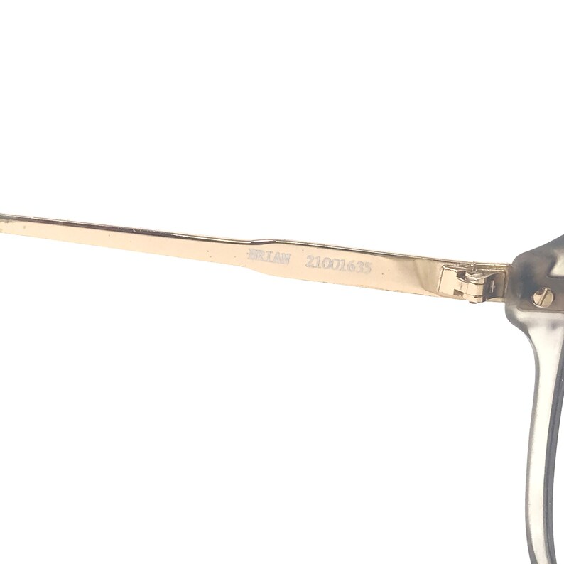 May include: Close-up of eyeglasses with a gold-colored arm. The text "BRIAN 21001635" is printed on the arm. The frame is a clear, translucent color.
