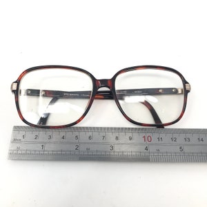 May include: Brown tortoiseshell eyeglasses with a rectangular frame. The glasses have clear lenses and silver accents on the temples. The text "SPECSAVERS 1764286" is visible on the frame. A ruler is placed below the glasses.