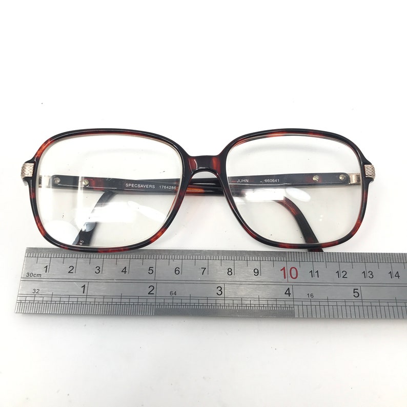 May include: Brown tortoiseshell eyeglasses with a rectangular frame. The glasses have clear lenses and silver accents on the temples. The text "SPECSAVERS 1764286" is visible on the frame. A ruler is placed below the glasses.