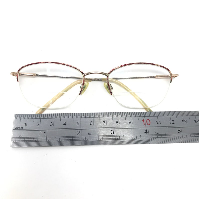 May include: Gold-tone half-rim eyeglasses with clear lenses. The top rims have a red and white speckled pattern. A metal ruler shows the glasses are approximately 5 inches wide.