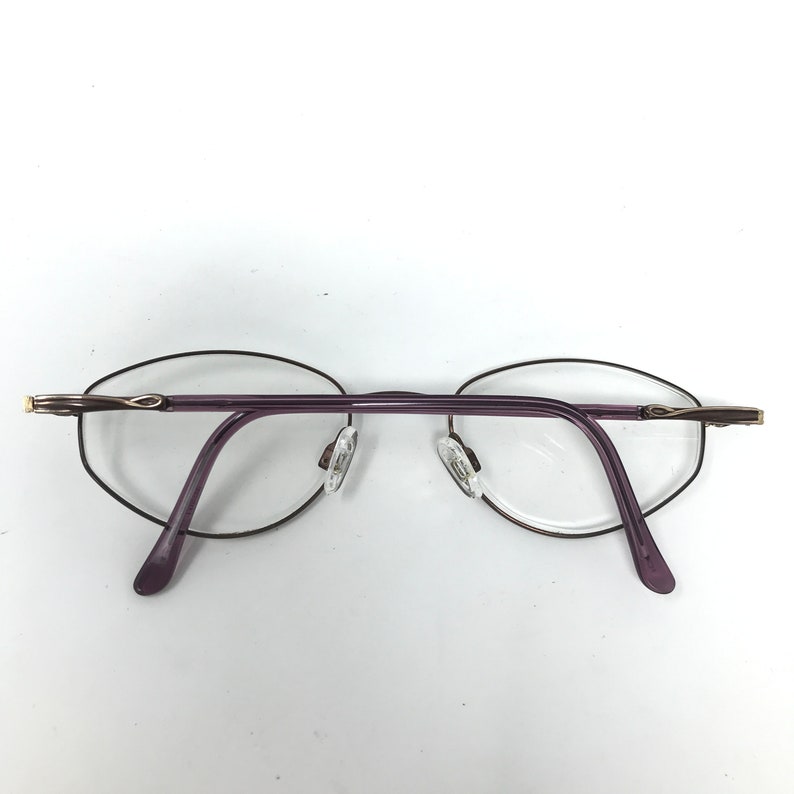 Puede incluir: Gafas sin montura con lentes ovaladas y montura morada. Las varillas son de color morado oscuro y las almohadillas nasales son transparentes. La montura tiene detalles dorados en las patillas. Las gafas est&aacute;n sobre un fondo blanco.